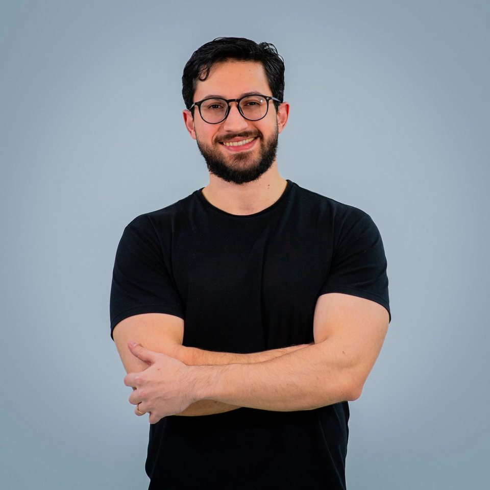 Retrato de Natan Klein, de óculos e camiseta preta, sorrindo com os braços cruzados